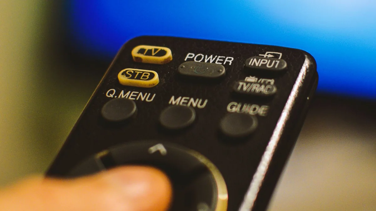 A hand holding a television remote in front of a blurred screen