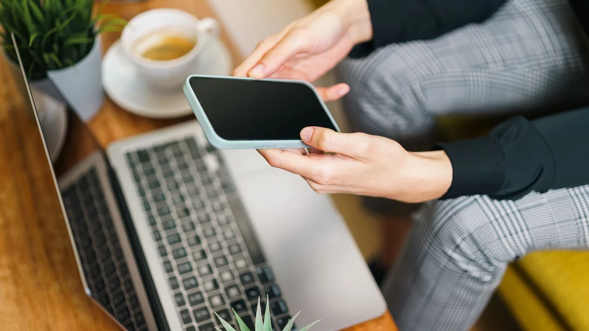 Hands holding a smartphone above a laptop on a desk